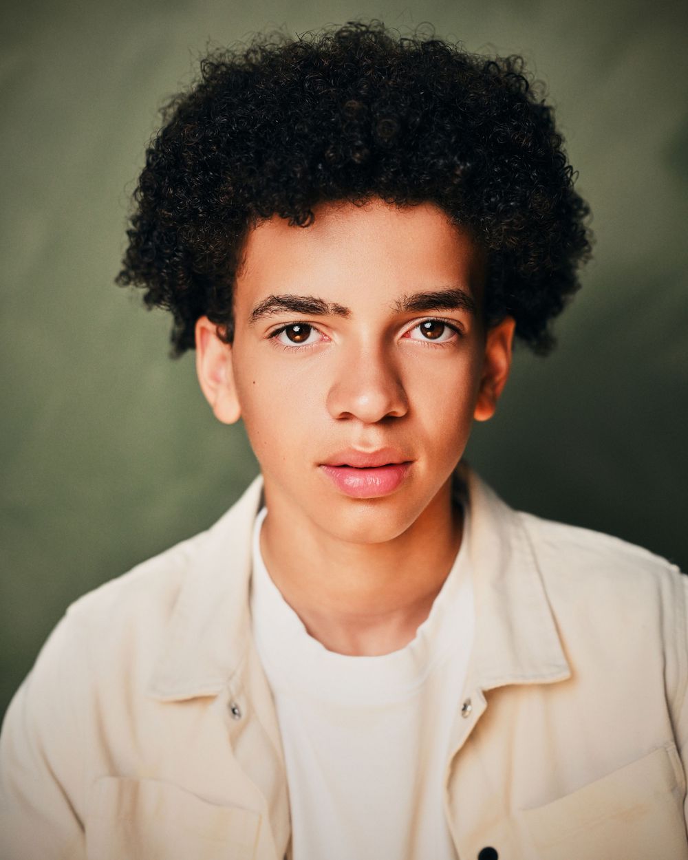 Young man with curly black hair wearing a cream jacket, posing for a professional headshot portrait.
