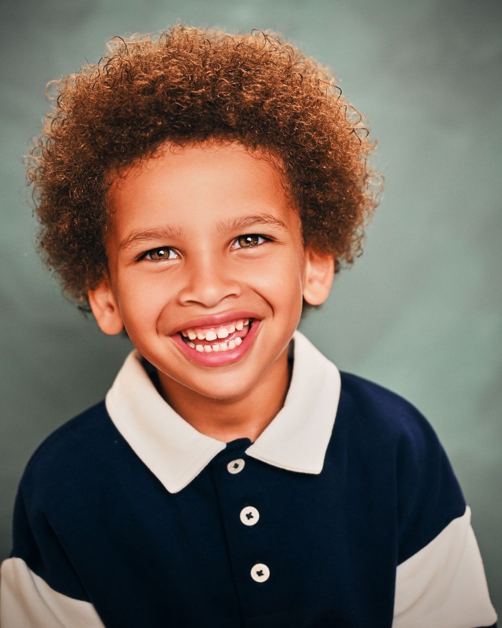 Smiling young boy with curly auburn hair wearing a navy and white polo shirt against a gray background.