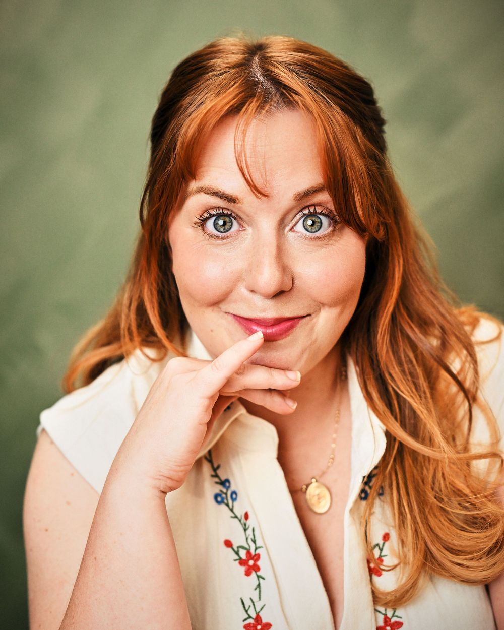 Smiling redhead woman with blue eyes posing playfully with finger on chin, wearing floral blouse and gold necklace.