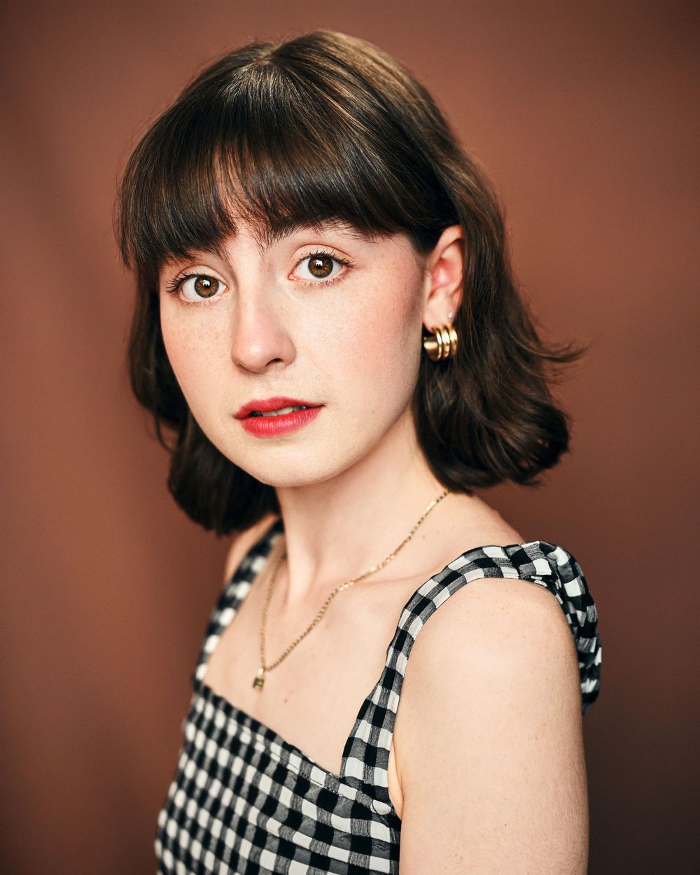 Young woman with short dark hair, bangs, gold hoop earrings, and a gingham top poses against a brown background.