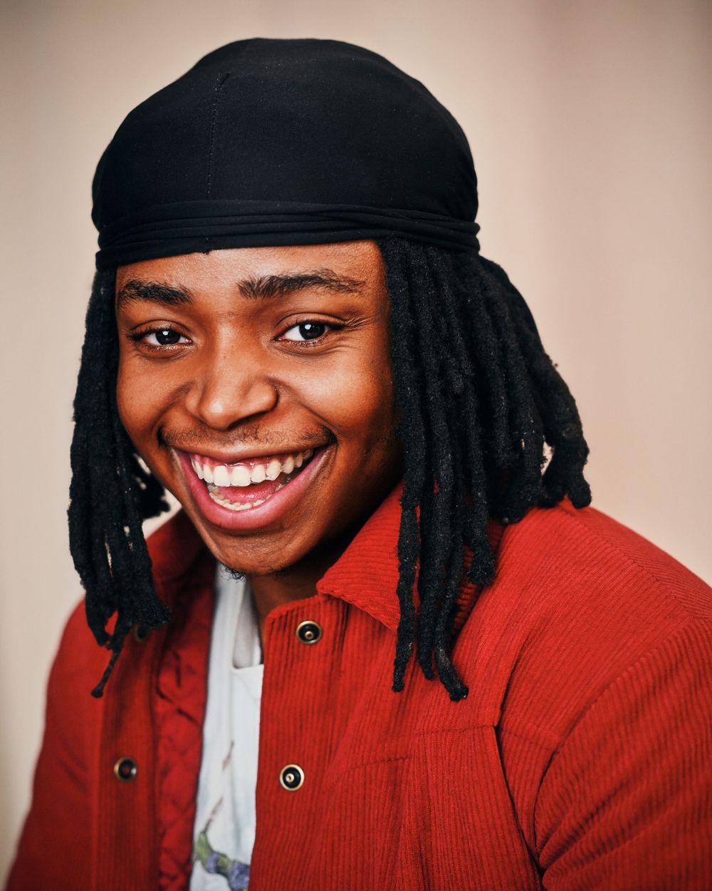 Smiling young man with dreadlocks and black durag wearing a red corduroy shirt against a neutral background.