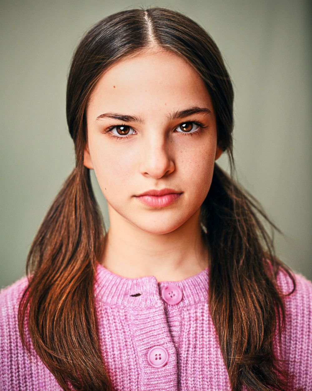 Young girl with dark hair in pigtails wearing a pink knit cardigan, posing for a professional headshot photo.