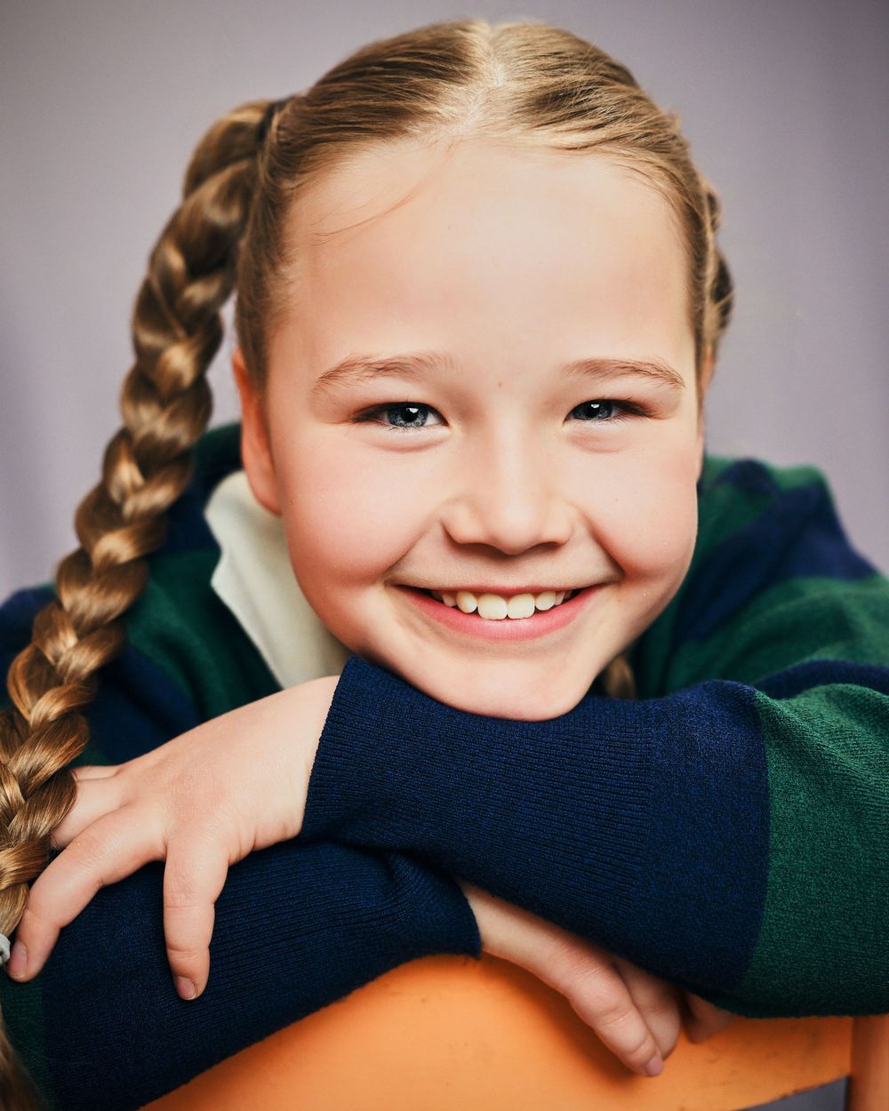 Smiling young girl with braided pigtails wearing a green and navy school uniform, leaning on a desk.