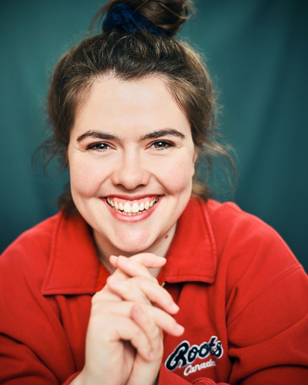Smiling young woman with hair bun wearing a red Roots Canada sweatshirt, posing with clasped hands against teal background.