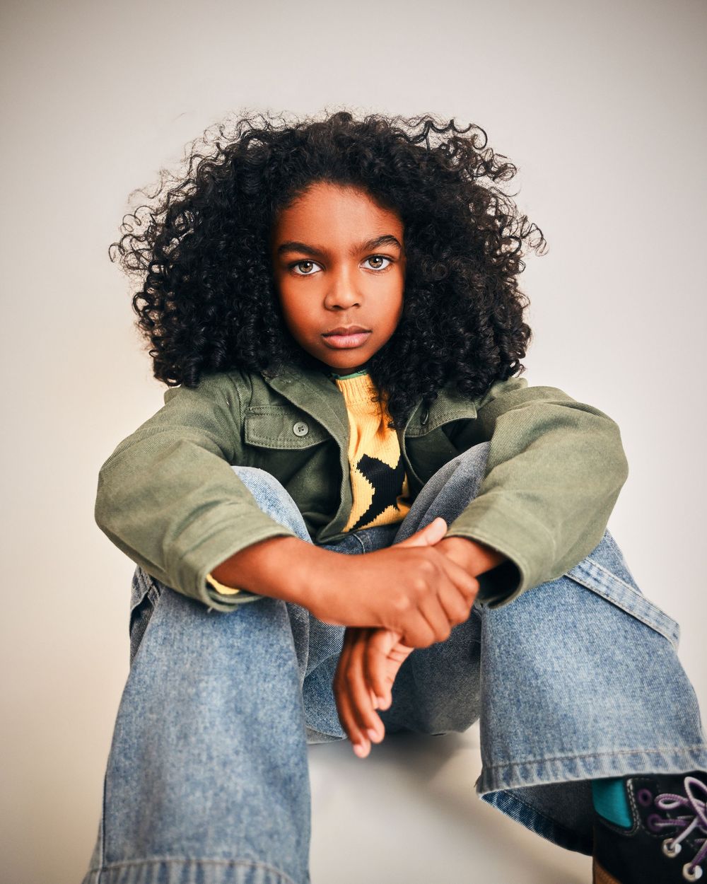 Young child with curly hair wearing a green jacket and jeans, sitting in a relaxed pose against a light background.