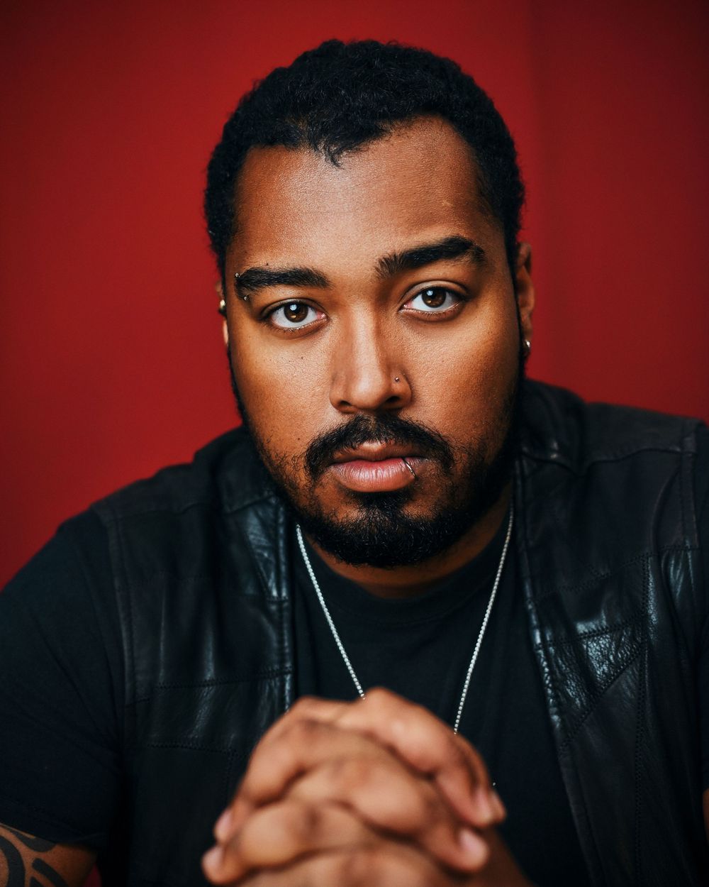 A serious young Black man with a beard poses with clasped hands against a bold red background.