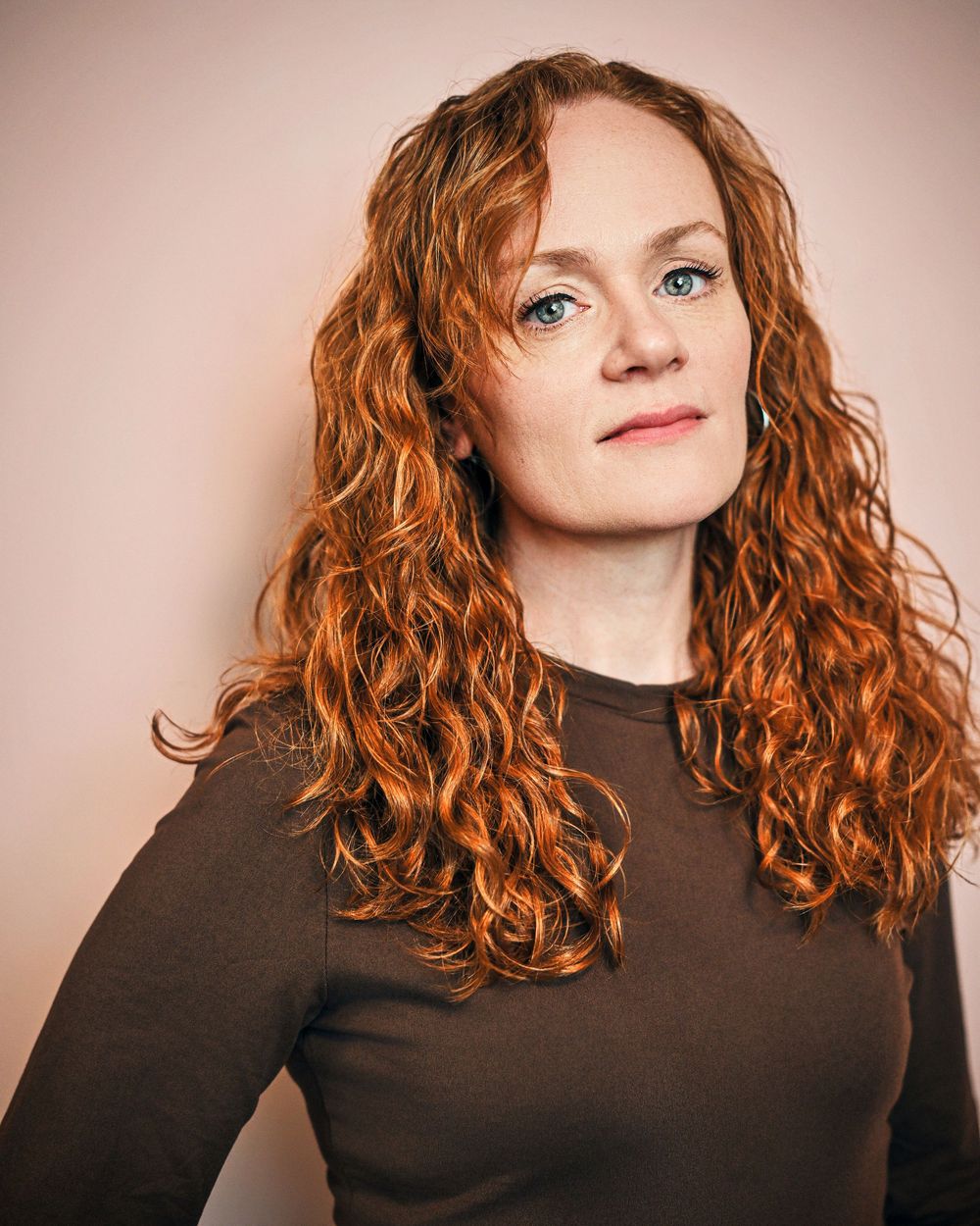 Woman with curly red hair and blue eyes wearing a dark brown top poses for a professional portrait.