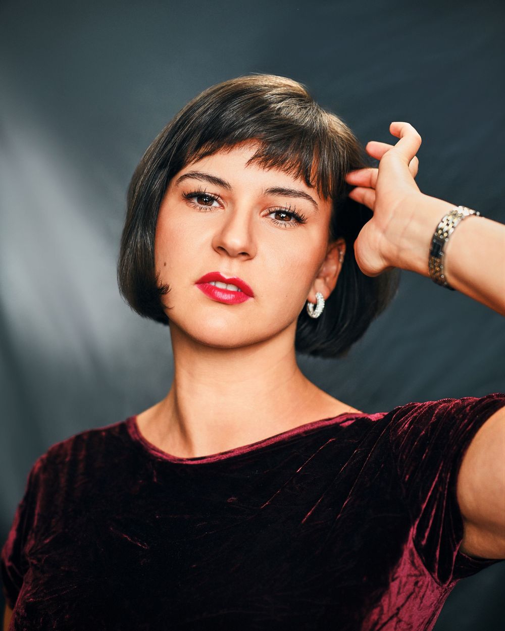 Woman with short bob haircut and red lips wearing a burgundy velvet top, posing confidently in a studio.