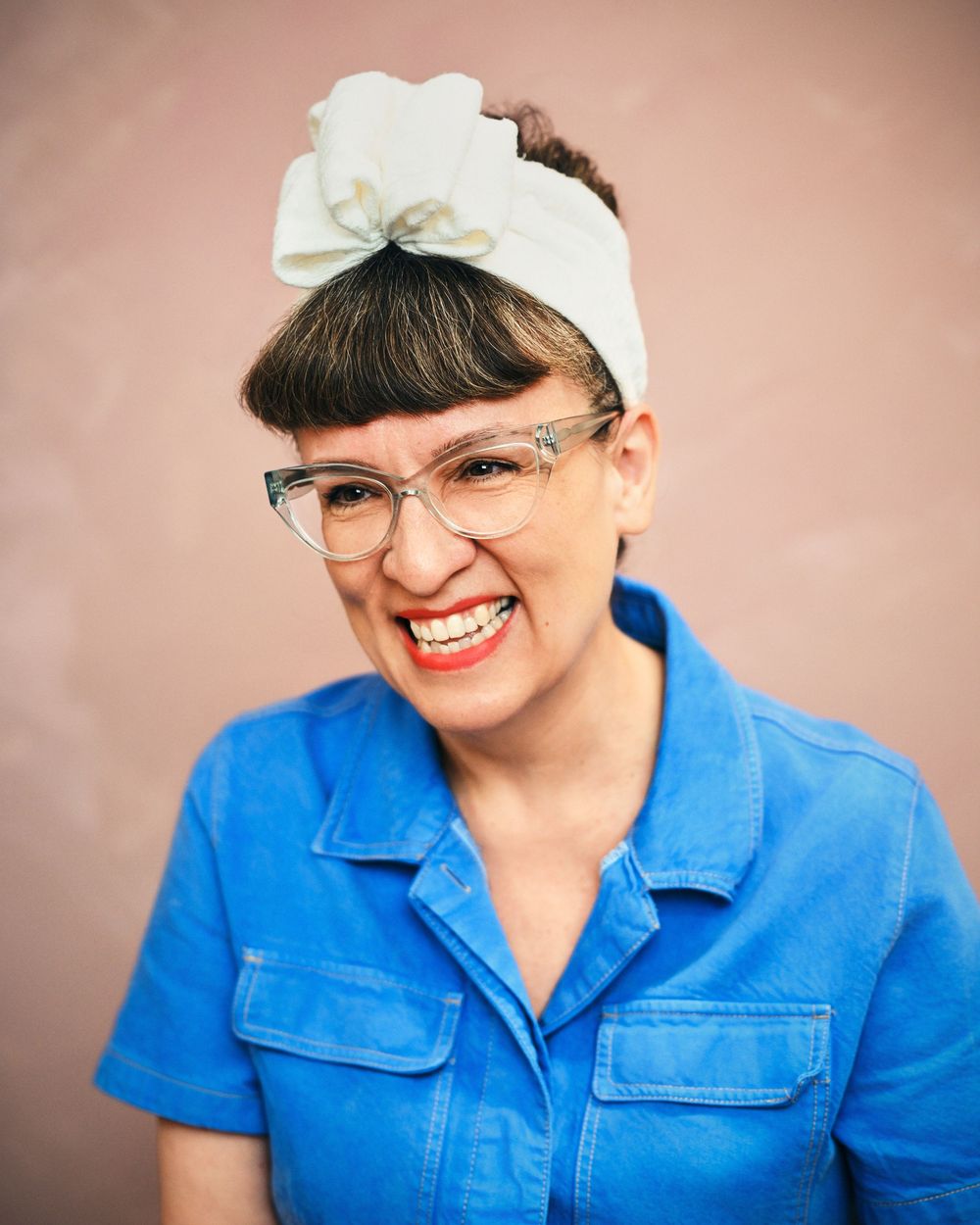 Smiling woman with bangs, clear glasses, white bow headband, and blue denim shirt poses against a pink background.