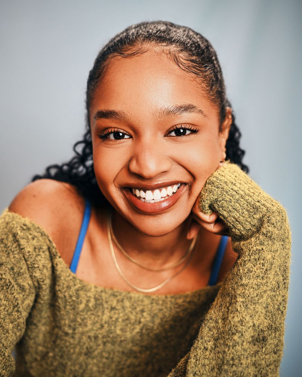 Young woman with curly hair smiling brightly, wearing a olive green sweater, in a professional headshot photo.