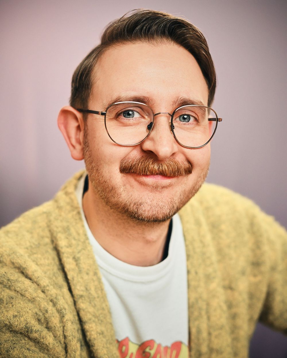 Smiling man with mustache, round glasses, and yellow cardigan poses for a professional headshot photo.