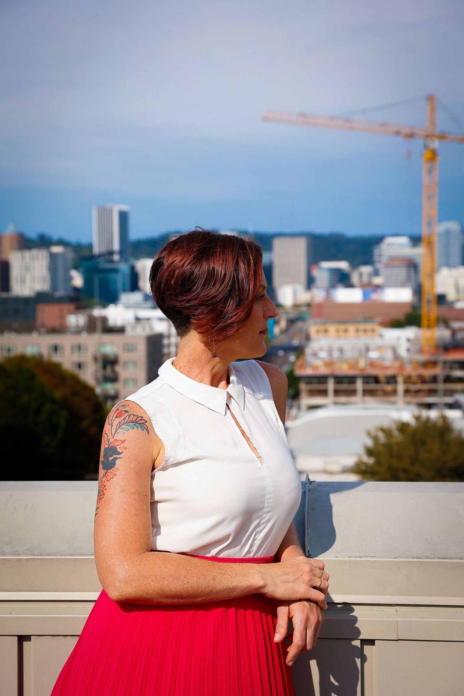 Person in white top and red skirt stands on rooftop with city skyline and construction crane in background.