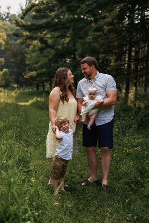 Family enjoys tender moments together in a lush green meadow surrounded by forest during golden hour photography session.