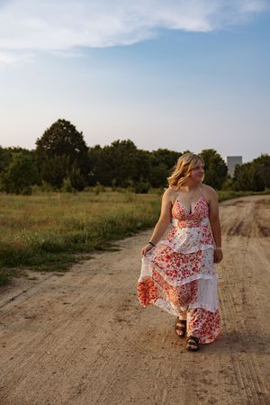 Someone wearing a flowing red and white floral dress walks along a dirt path during golden hour in a rural setting.