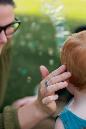 A close-up view shows hands gently touching a child's ear and neck area against a blurred green background.