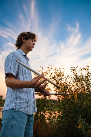 A person in a blue plaid shirt holds a shovel at sunset in a garden with silhouetted trees in the background.
