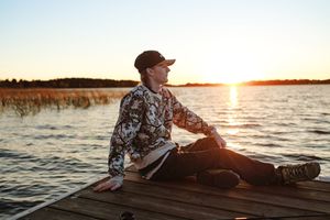 Person sitting on a wooden dock at sunset by a calm lake, wearing a floral jacket and cap, enjoying the scenic view.
