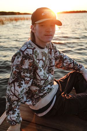 Teen boy in floral camo sweatshirt and cap sits on a boat dock at golden sunset over calm water.
