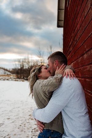 Couple sharing a romantic kiss against a red barn wall in a snowy winter landscape at sunset.