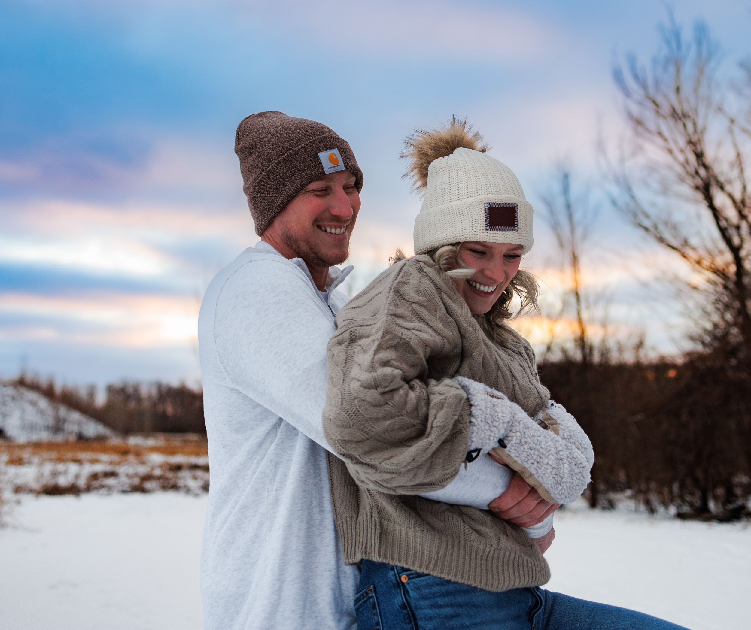 Couple laughing together in snowy winter landscape wearing matching beanies at sunset.