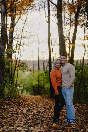 A couple poses on an autumn forest trail covered in fallen leaves at sunset.