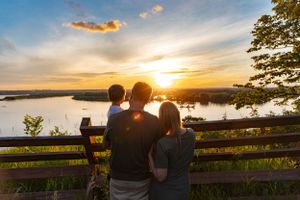 Silhouettes watching sunset over lake from scenic overlook with wooden fence.