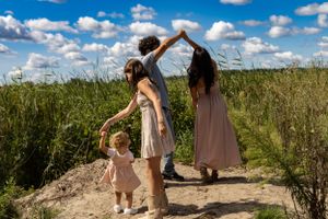 Three people dance and play with a small child on a dirt path through tall grass.