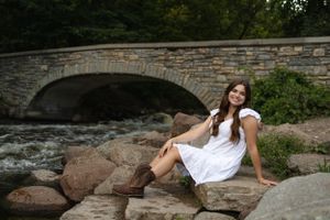 Young woman in white dress posing on rocky stones near a stone bridge.