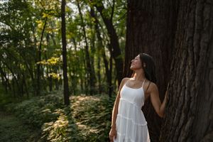 Person in white dress leaning against large tree trunk in sunlit forest.