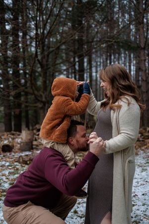 Family poses together in a snowy forest setting with a small child in a brown bear coat reaching upward.