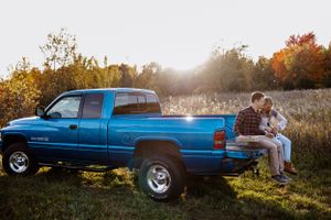 Couple sitting on tailgate of blue pickup truck during golden hour in rural setting.