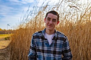Young man in plaid flannel shirt standing among tall wheat grass under blue sky.