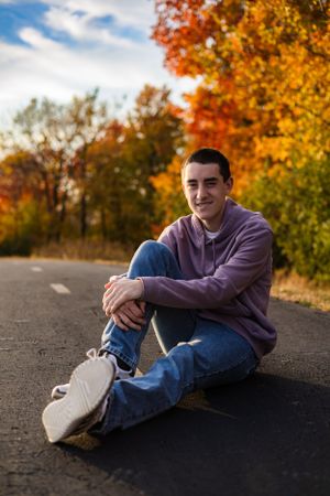 Casual portrait of young man sitting on road with autumn foliage in background.