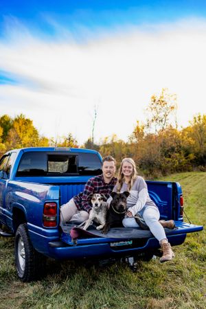 Couple and their dog pose in the bed of a blue pickup truck on sunny autumn day.