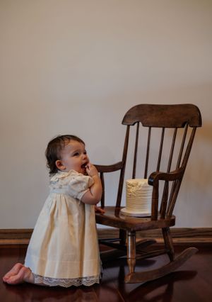 A baby in a white lace dress examining a birthday cake on a wooden chair from different angles in a photo sequence.