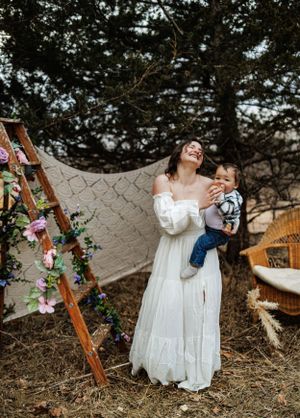 A rustic outdoor scene with a white dress, decorated wooden ladder, and macrame backdrop against evergreen trees.