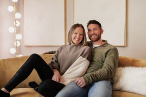 Happy couple cuddling together on a tan couch with string lights and artwork in the background.