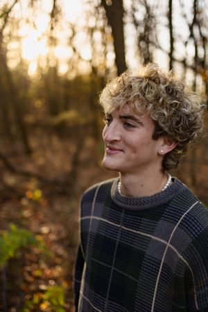 Person in dark plaid sweater smiling against autumn forest backdrop during golden hour.