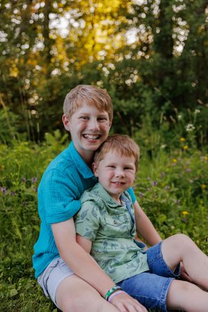 Two young children sitting together and smiling outdoors in a natural setting with golden sunlight and greenery.