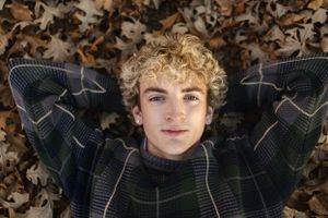 Person with curly blonde hair lying on autumn leaves wearing a plaid sweater looking up at camera.