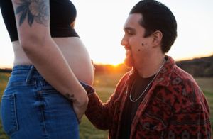 A pregnant belly is gently held against a scenic sunset background as orange light glows in a rural setting.