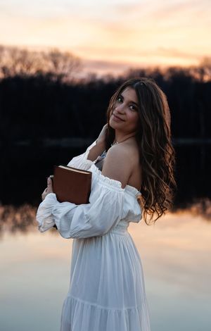 Woman of faith in flowing white dress poses artistically by lake during sunset.