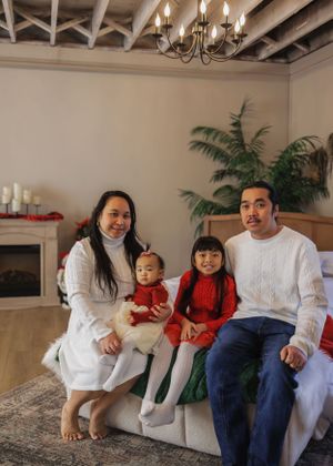 Family of four in white sweaters sits together on couch by fireplace during holiday season.