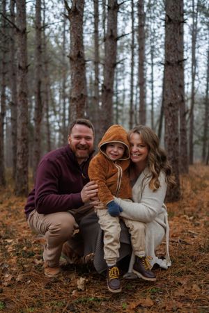 Family of three posing together in an autumn forest with child wearing cozy brown hoodie.