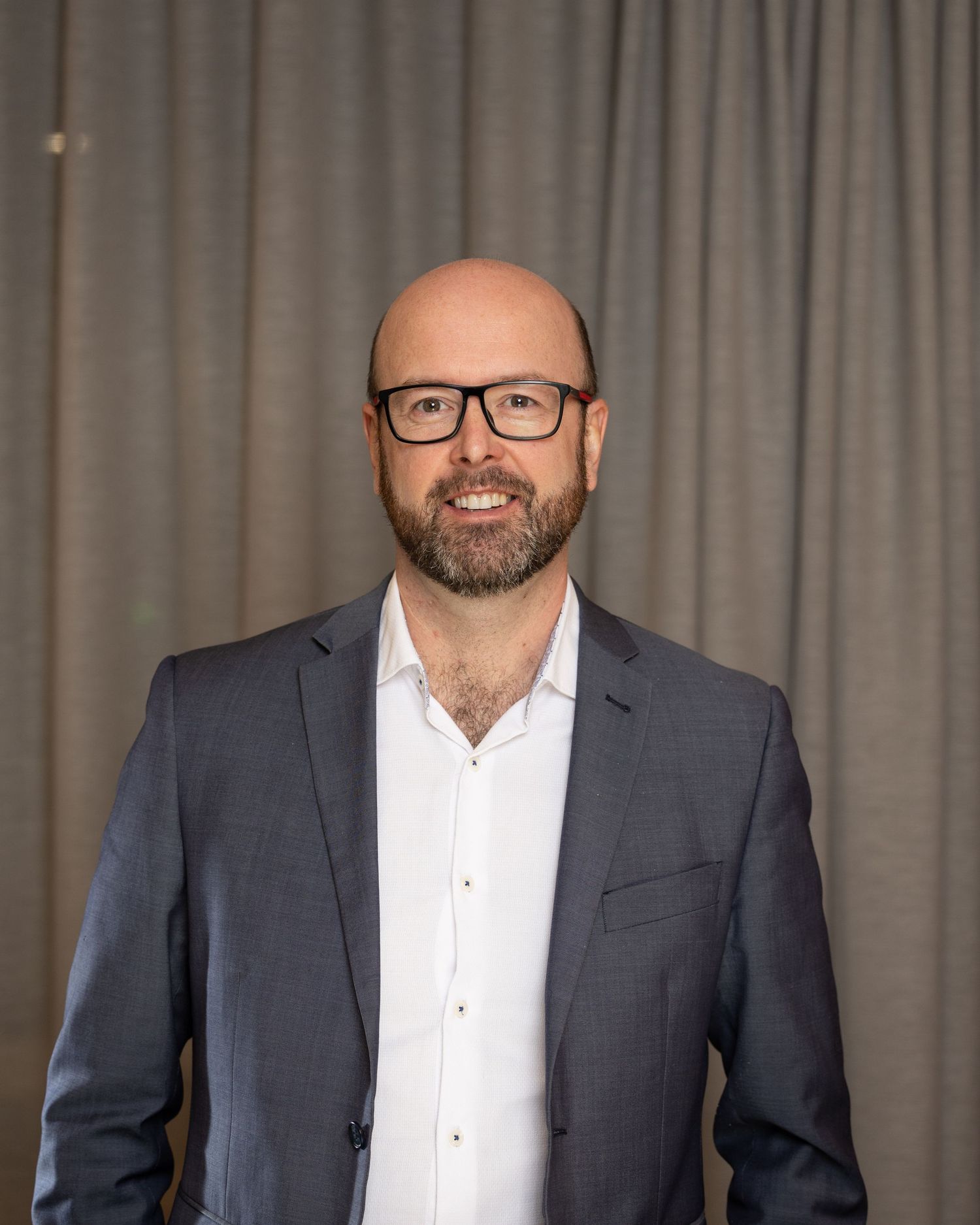Professional headshot of a bearded man in glasses wearing a grey suit jacket and white shirt.