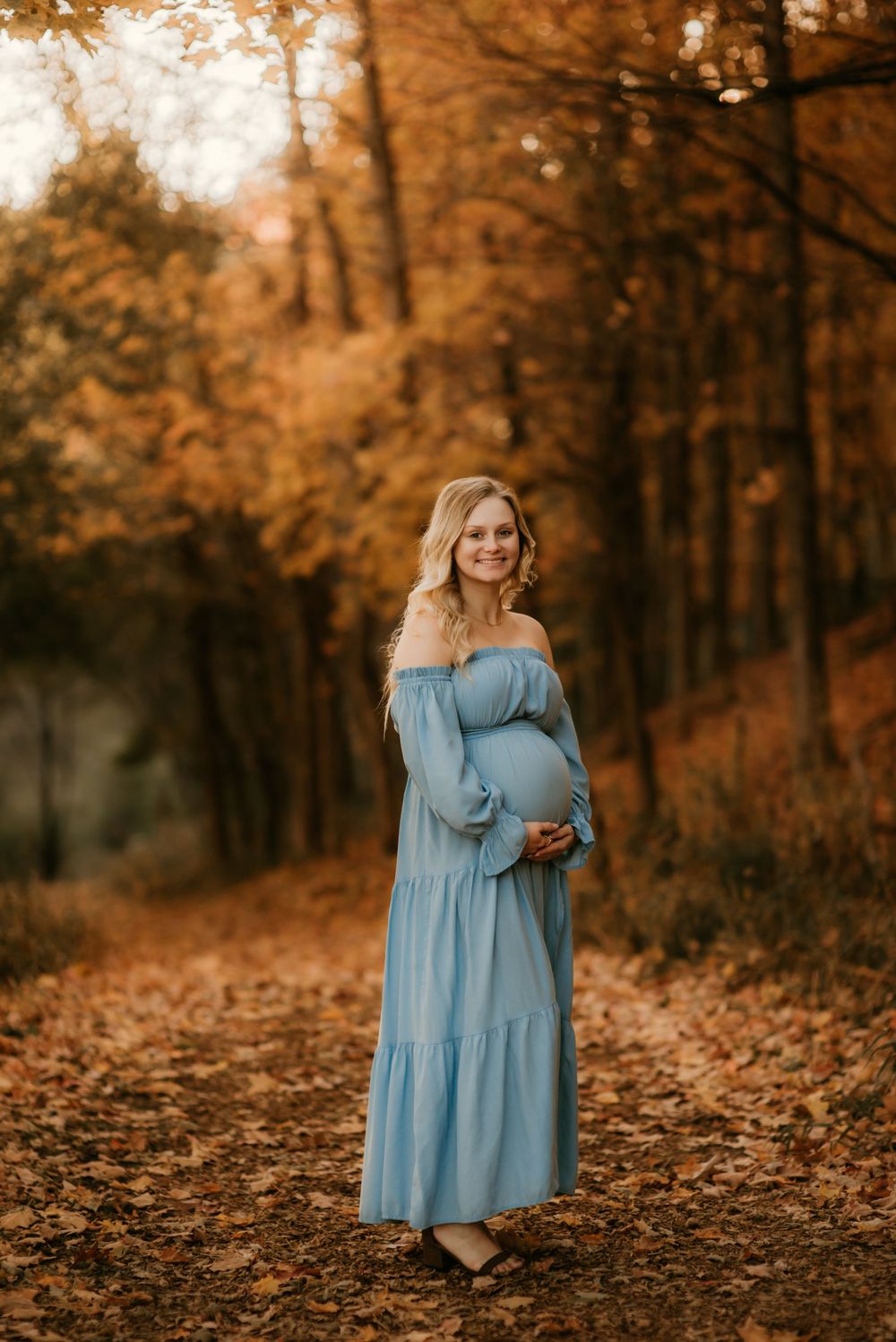 Person in light blue dress stands among fallen autumn leaves in a forest with warm orange tones. maternity session inspo