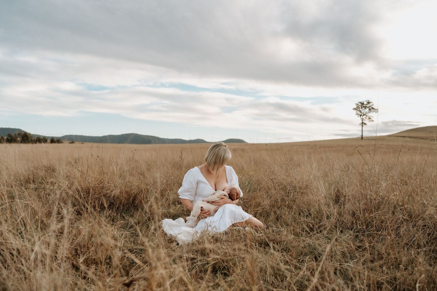 Woman in white dress sitting in golden field, breastfeeding newborn baby at sunset.