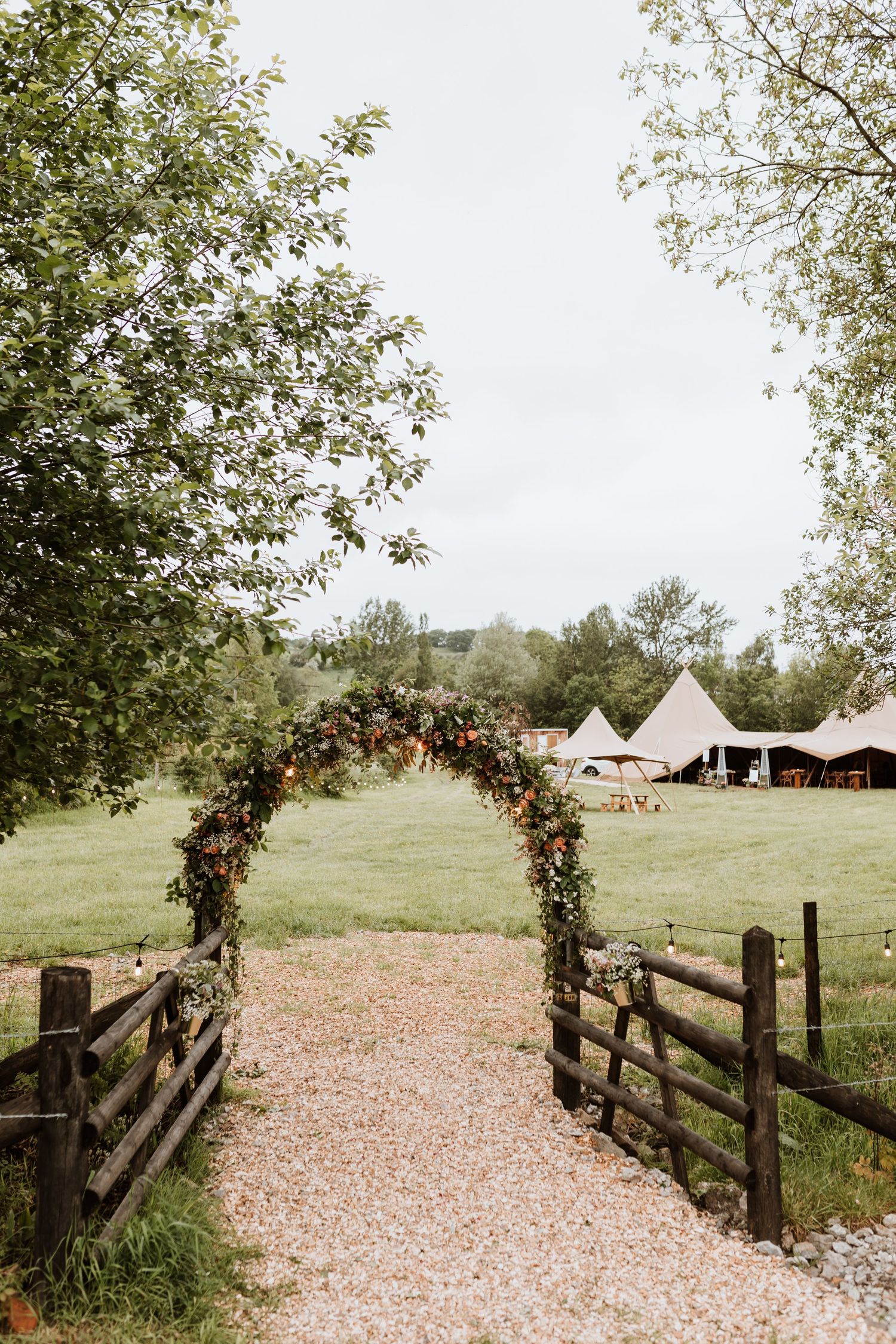 A rustic floral arch over a gravel path leads to bell tents and a meadow, creating a romantic outdoor wedding setting.