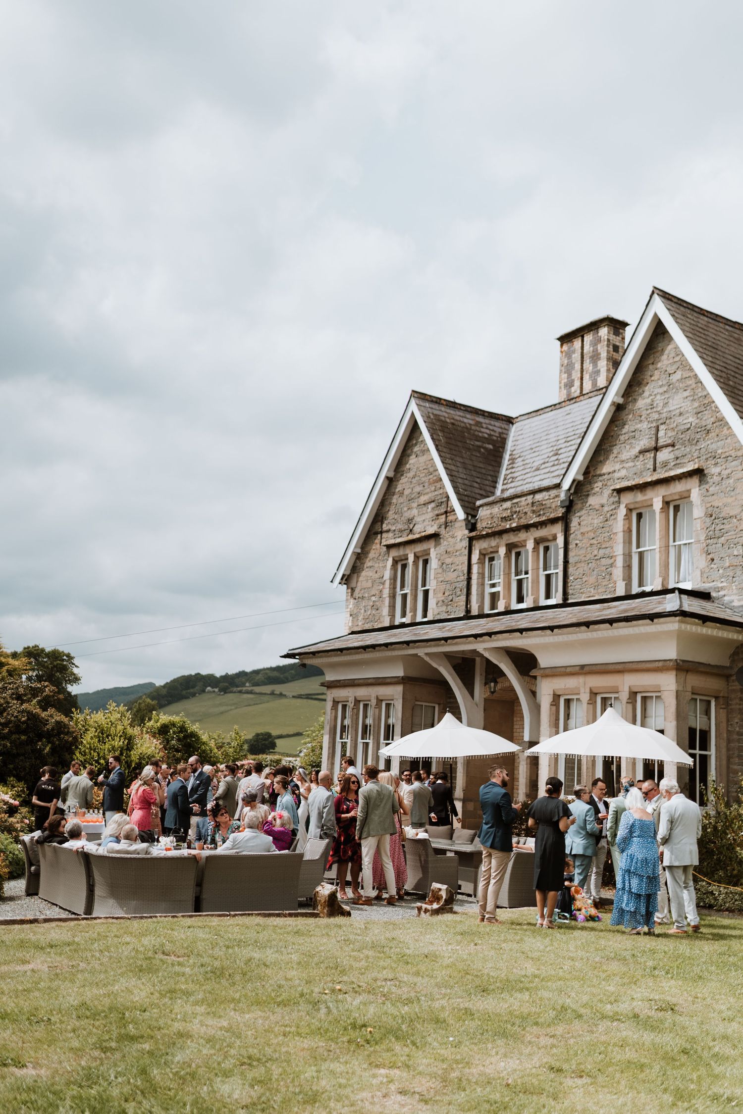 Elegant countryside manor house hosting an outdoor garden party with guests gathering on the lawn under cloudy skies.