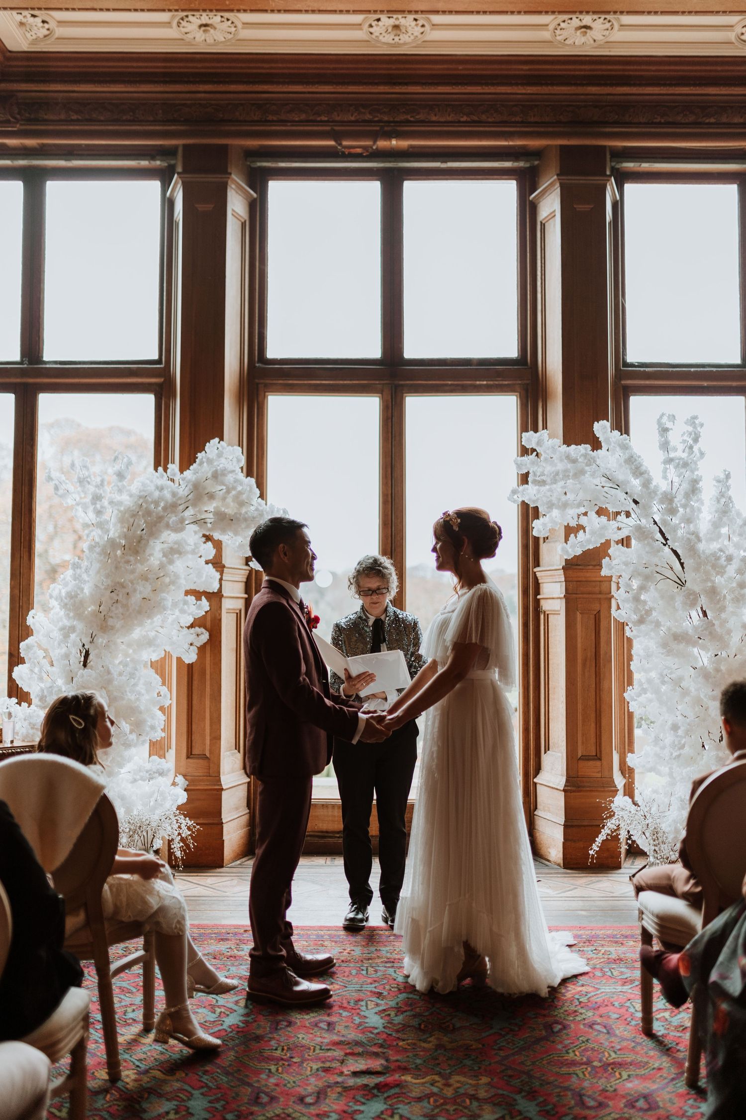 A romantic winter wedding ceremony in an elegant room with ornate windows and white floral arches on a patterned carpet.